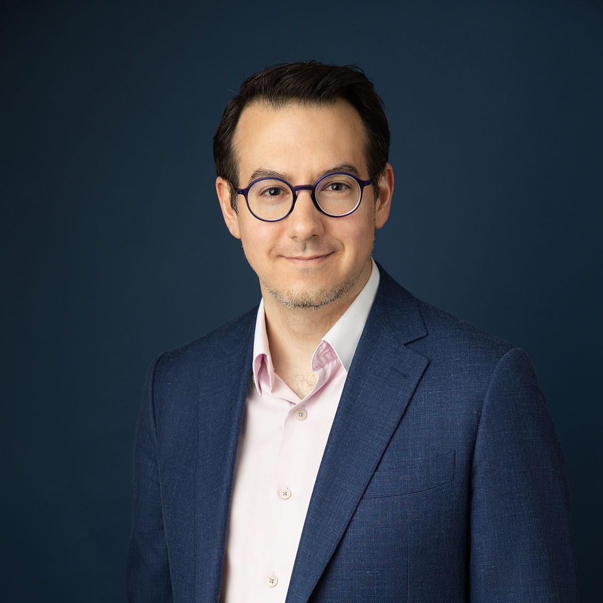 Un homme aux cheveux noirs courts, portant des lunettes rondes, une chemise rose clair et un blazer bleu foncé, se tient devant un fond bleu foncé uni, souriant légèrement.
