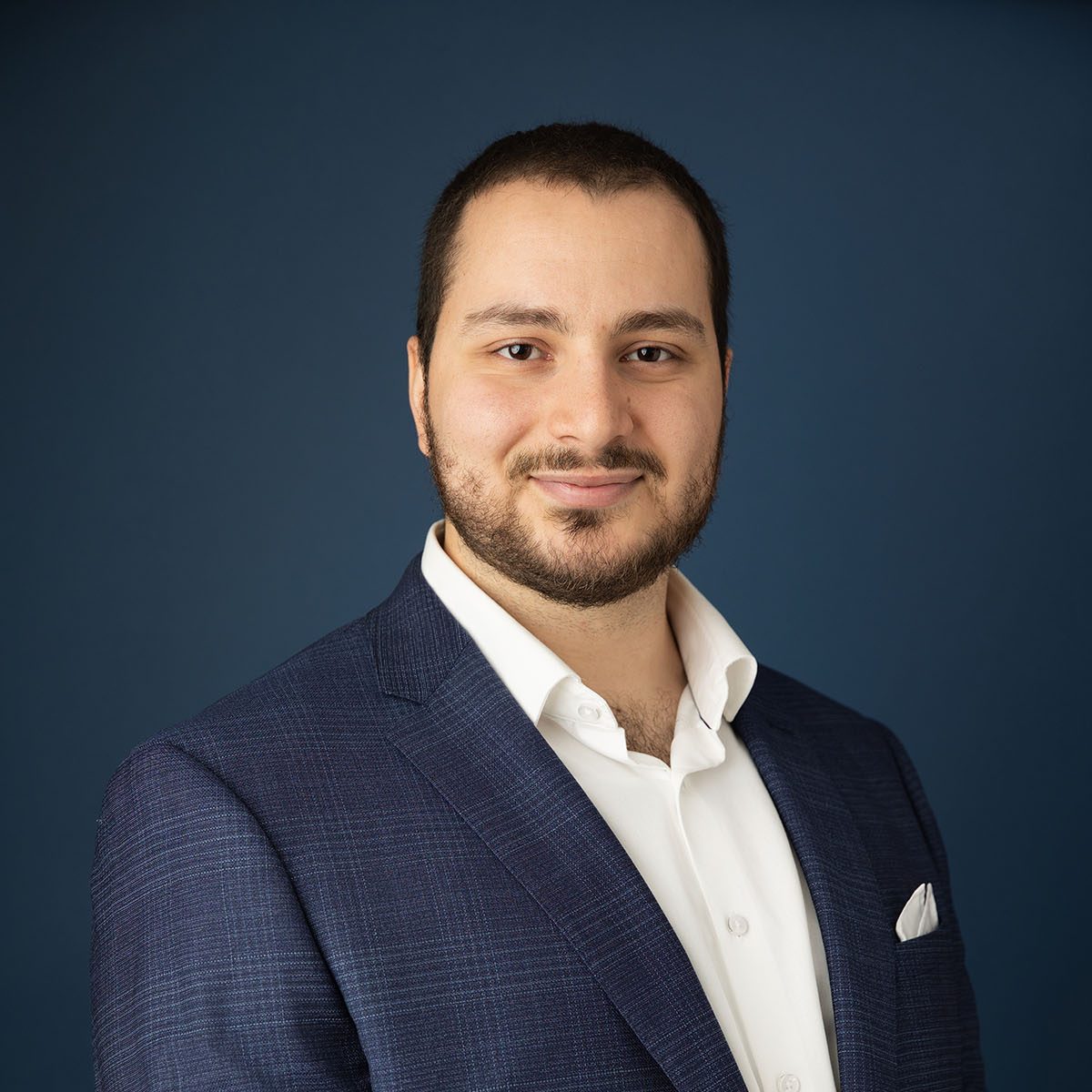 Un homme aux cheveux noirs courts et à la barbe taillée est vêtu d'une chemise blanche et d'une veste de costume bleu foncé. Il se tient debout devant un fond bleu foncé, souriant légèrement à la caméra. Il porte une pochette blanche dans sa veste.