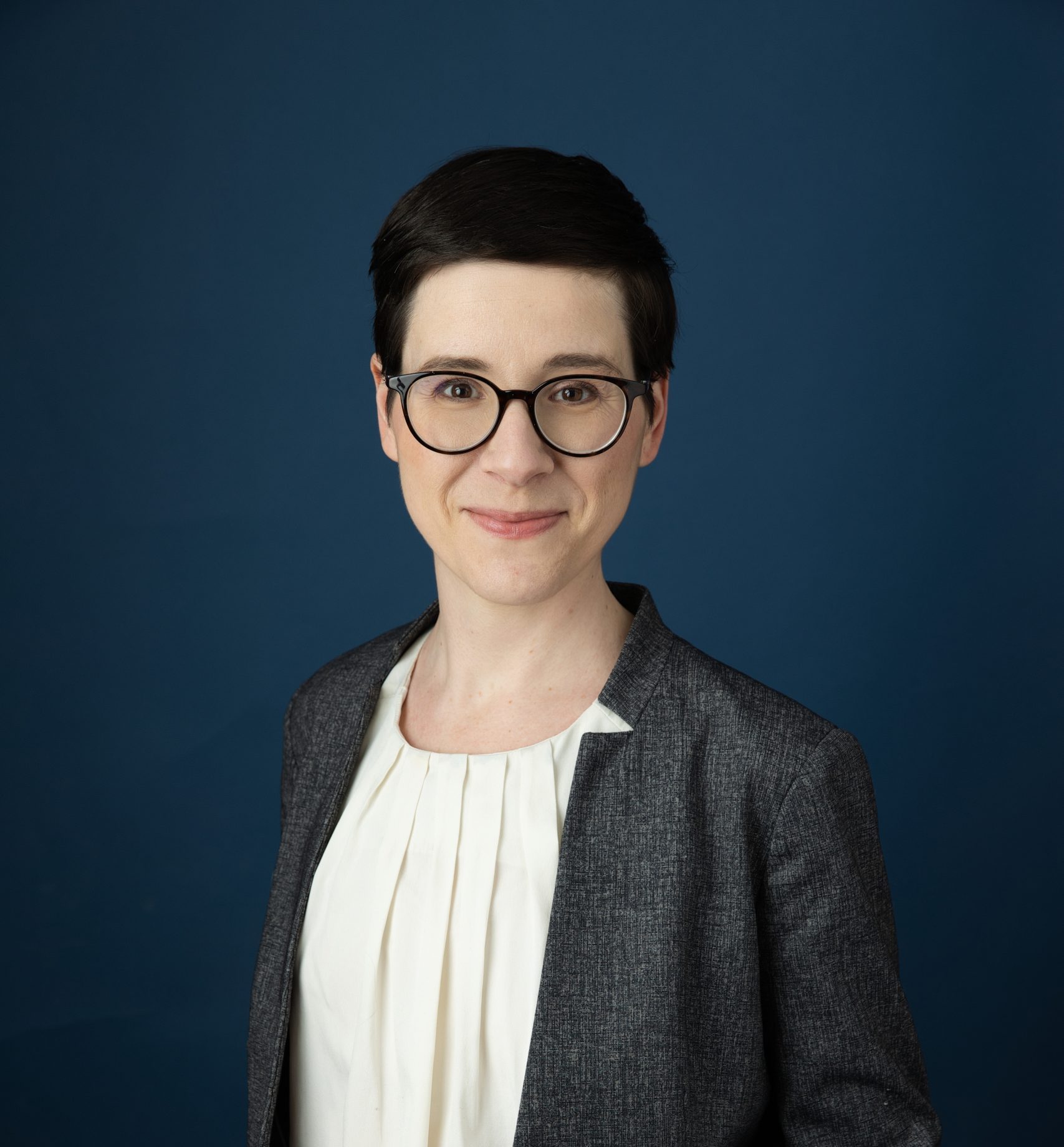 Evoto Femme aux cheveux bruns court portant des lunettes noires, un veston gris et une chemise blanche.